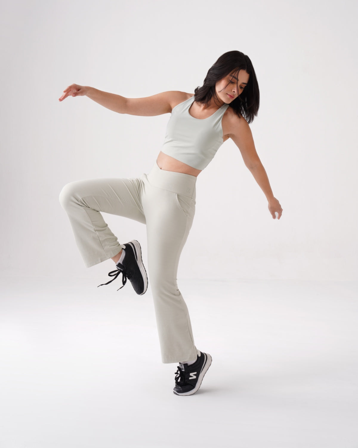 Woman in light beige RYZ Softretch® flared leggings and matching tank top posing mid-movement against a white studio background, showcasing flexible, no-front-seam activewear for women.